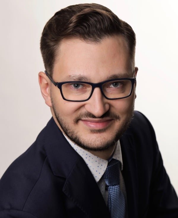 Zdjęcie portretowe mężczyzny w lekkim uśmiechu, brązowych włosach i okularach